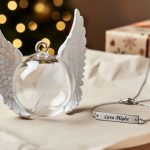 Memorial angel-wing and clear feather-ball ornaments placed with personalized bracelet and name-bar style jewelry on neutral holiday gift wrapping with blurred Christmas lights in the background.